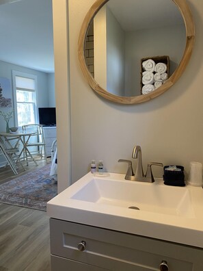 A bathroom with a white sink, a round mirror, and a wooden-framed mirror. There are toiletries on the sink, a towel rack, and a rug on the floor. - Hanscom's Motel and Cottages (Bar Harbor)