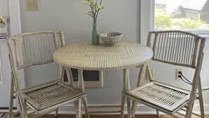 A dining area with a round table, two wicker chairs, a vase with flowers, and a painting of stacked stones on the wall. - Hanscom's Motel and Cottages (Bar Harbor)