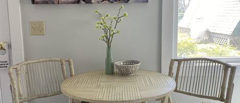 Ein Essbereich mit einem runden Tisch, zwei Korbstühlen, einer Blumenvase und einem Gemälde mit gestapelten Steinen an der Wand.