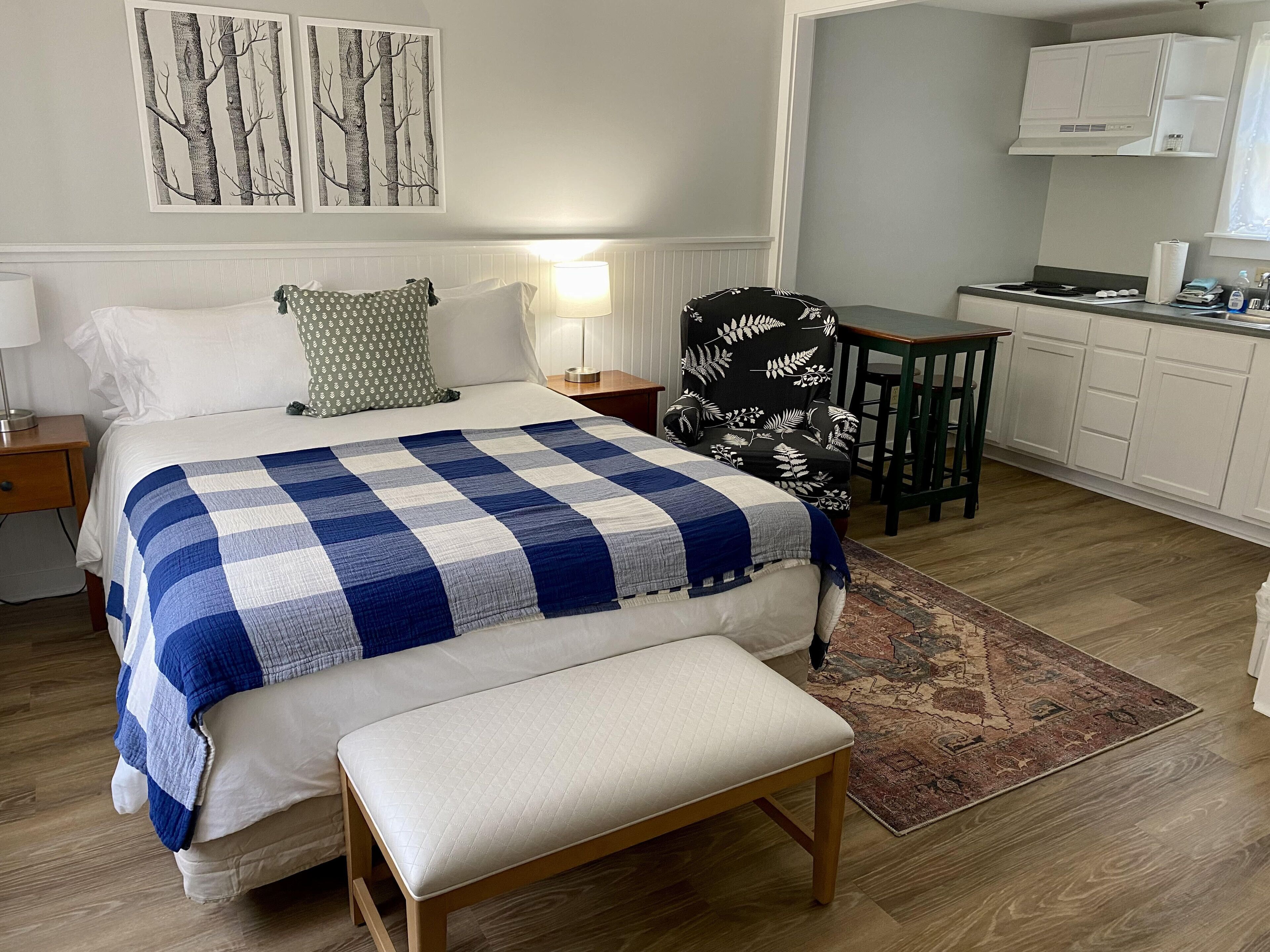 Quarto com cama, mesas de cabeceira, uma cadeira, um banco e uma área de cozinha com armários e bancada.