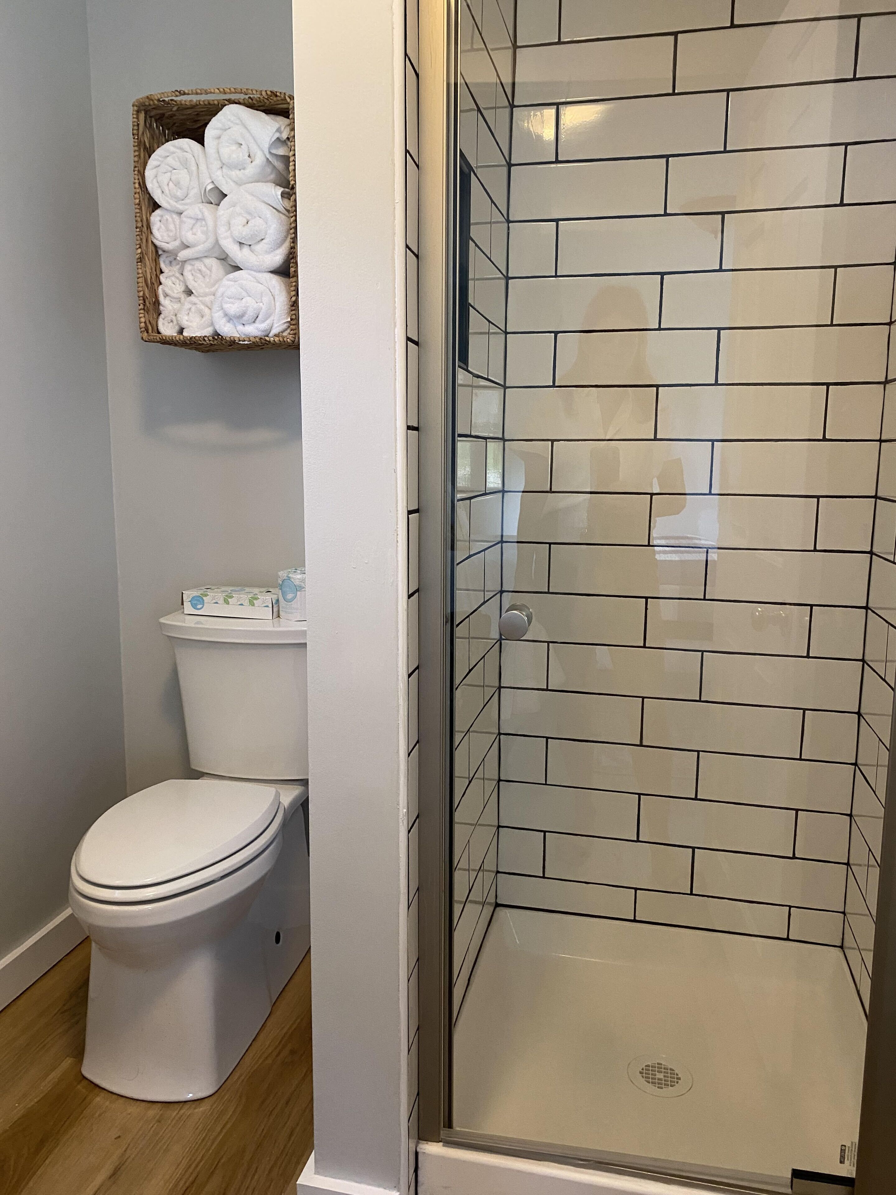 A bathroom with a toilet, a shower with tiled walls, and a basket of towels on the wall.