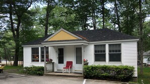 A small, single-story house with a porch, a red chair, and a yellow door. - Hanscom's Motel and Cottages (Bar Harbor)
