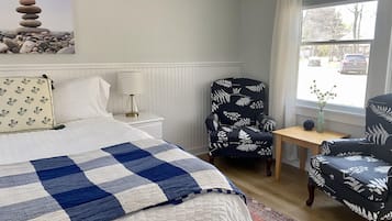 A neatly arranged bedroom with a bed, two chairs, a nightstand, a lamp, a window with a view of the street, and a painting on the wall.