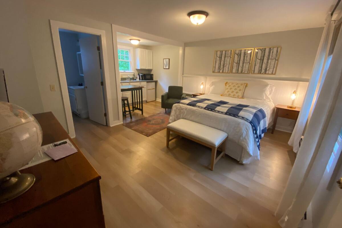 A bedroom with a bed, a bench, a desk, and a kitchenette in the background.