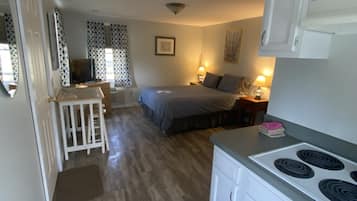 Una cocina funcional con armarios blancos y una estufa, un pequeño comedor con mesa y sillas, y un dormitorio con cama y mesita de noche.