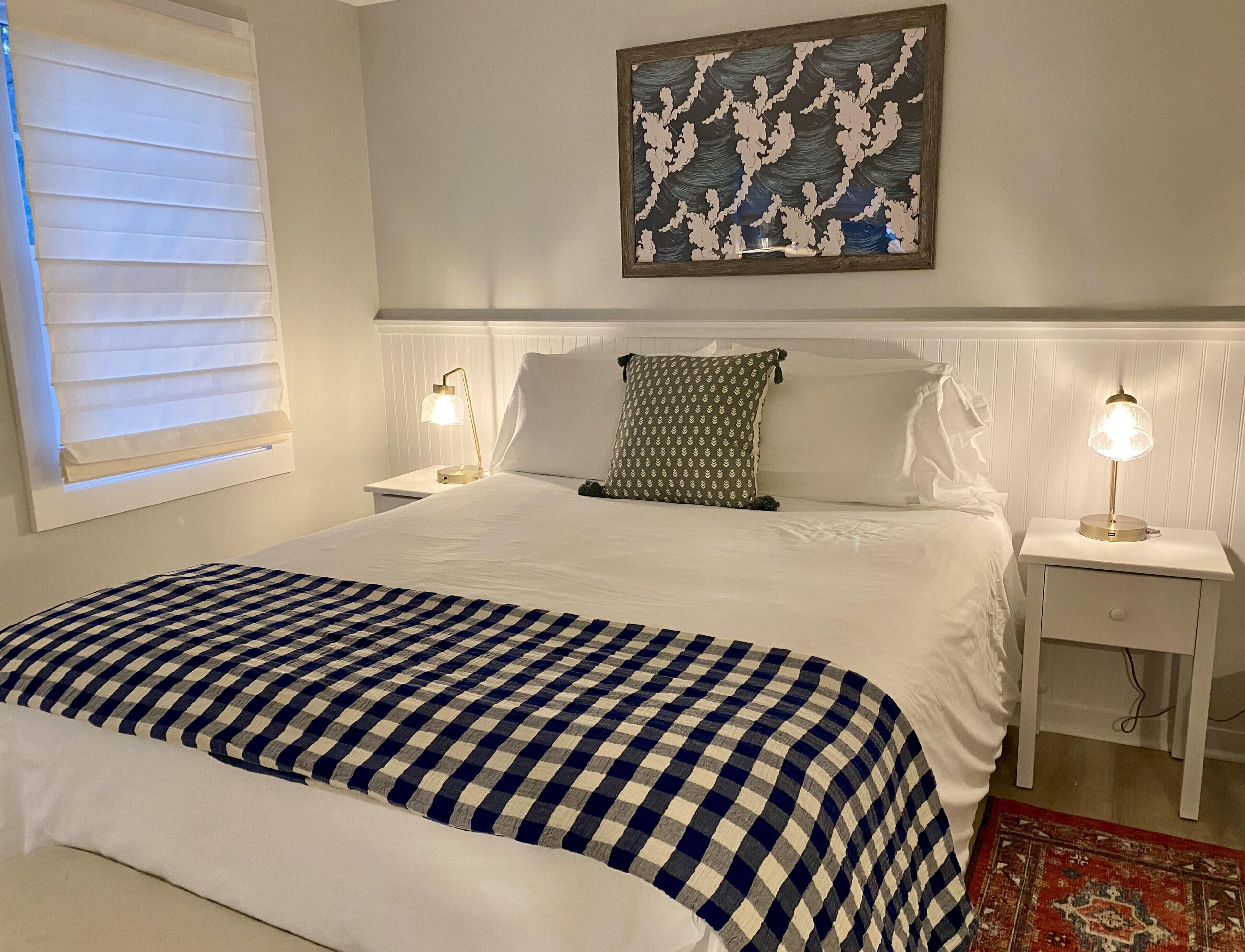 A neatly made bed with a checkered bedspread, two bedside lamps, a wall-mounted artwork, and a window with blinds.
