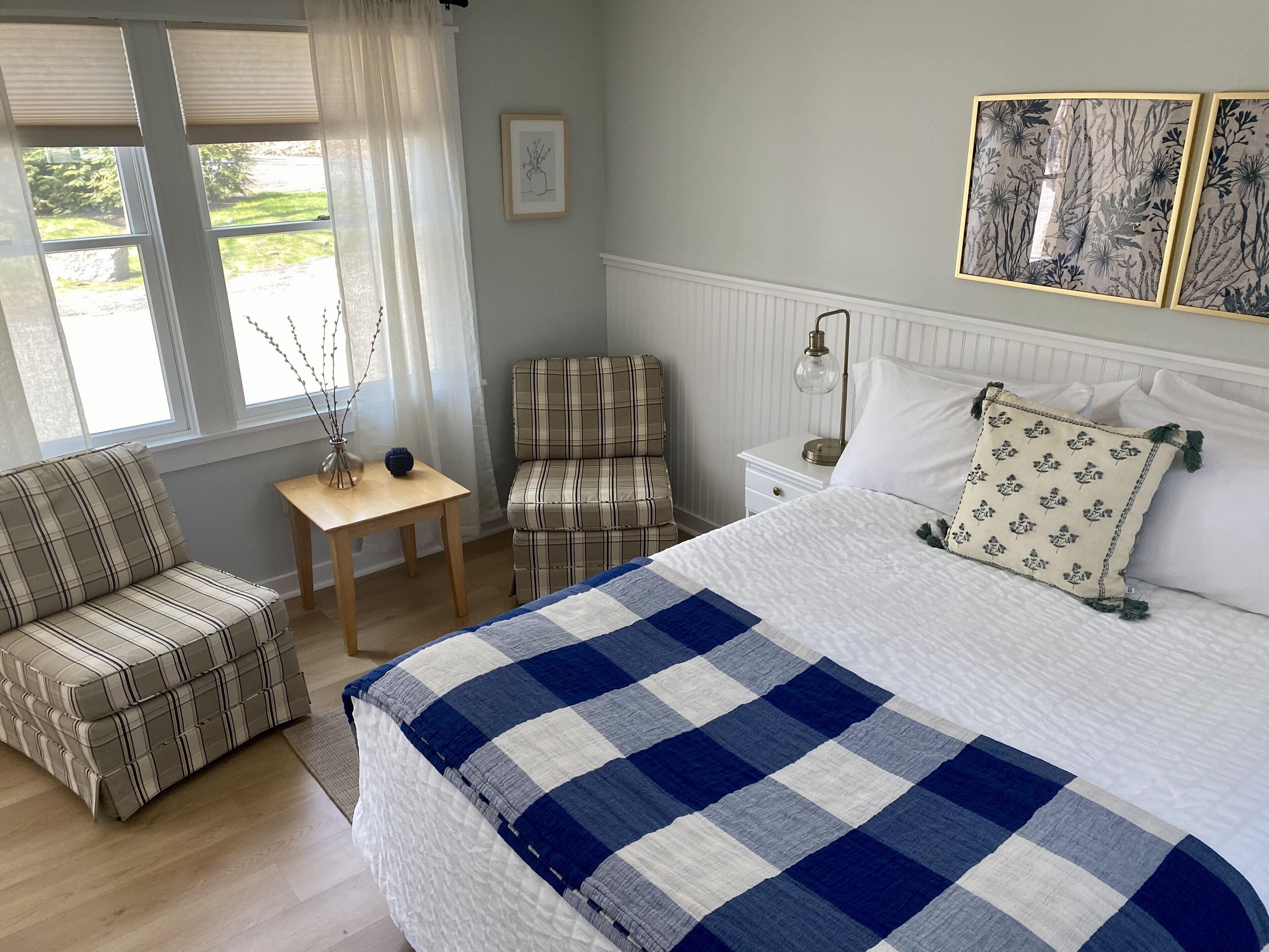 Une chambre à coucher avec un lit, une chaise, une table de chevet, une lampe et une fenêtre avec des rideaux.