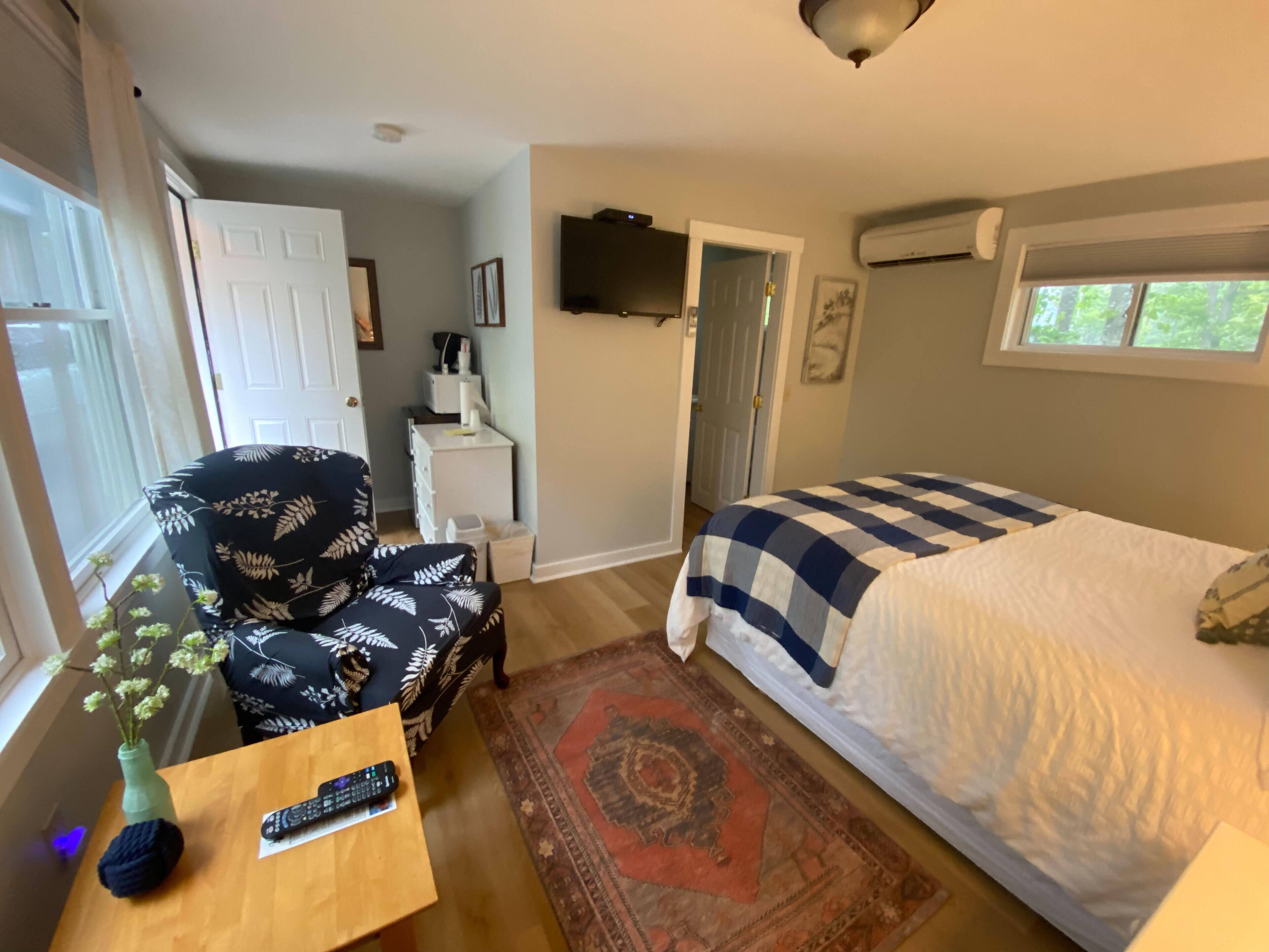 A bedroom with a bed, a chair, a small table with a remote control, a vase with flowers, and a TV on the wall.