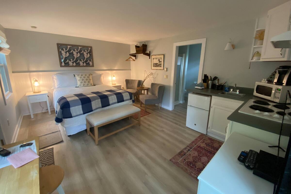 A compact hotel room with a bed, bedside table, chair, desk, kitchenette, and a small bathroom.