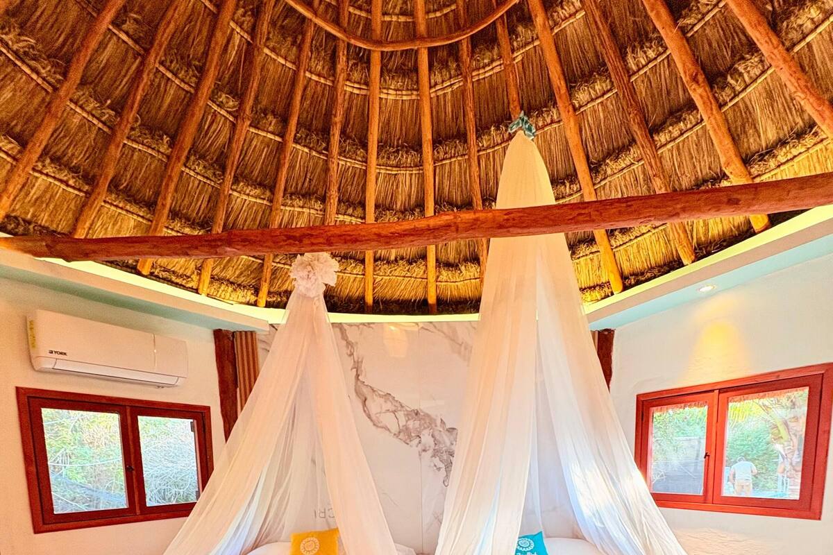 A room with a thatched roof, two beds with white canopies, wooden flooring, and windows with curtains.