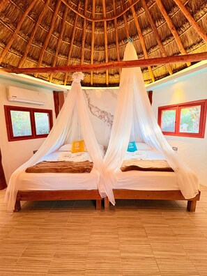 Una habitación con techo de paja, dos camas con dosel blanco, suelo de madera y ventanas con cortinas.