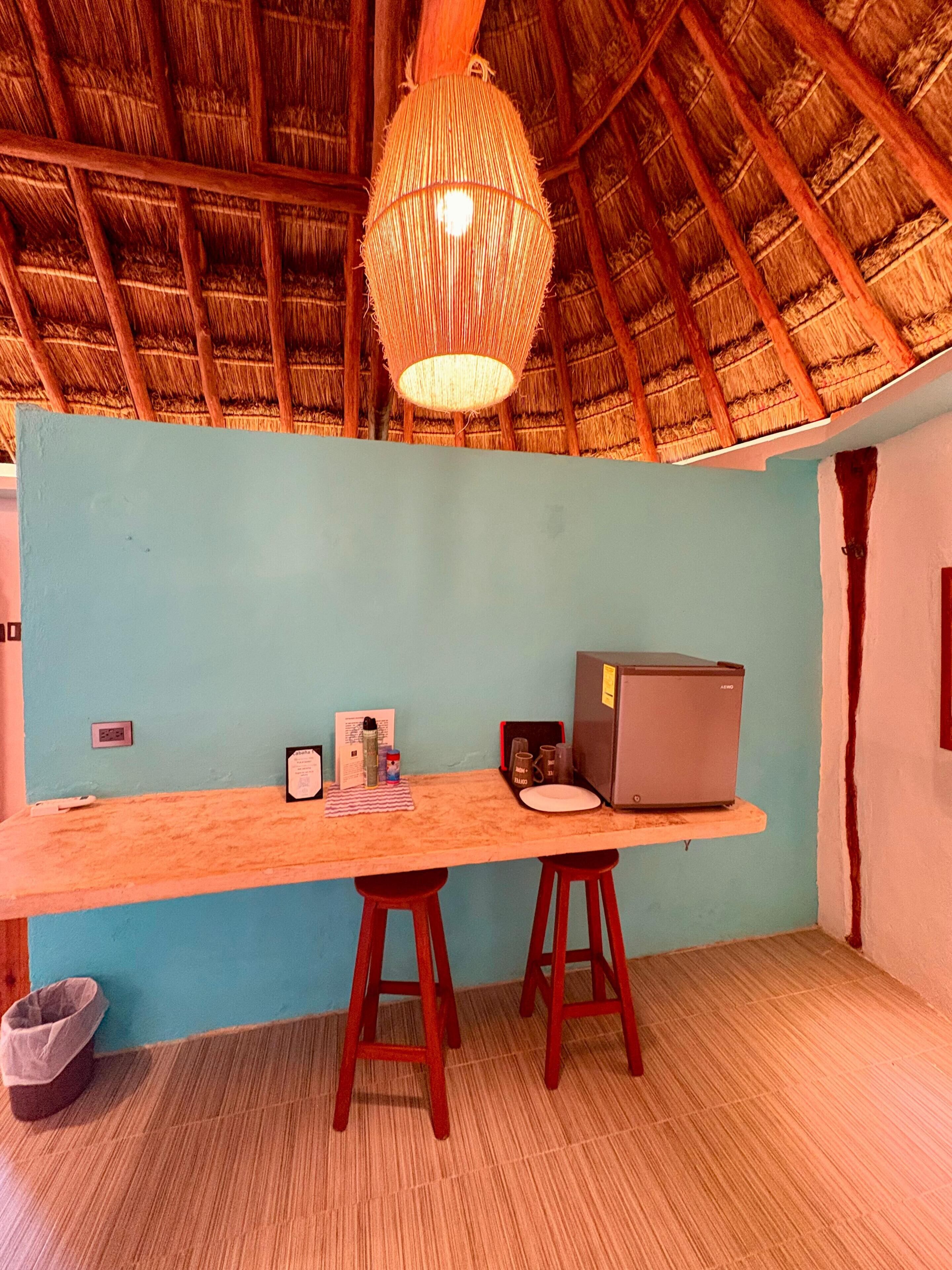A room with a long wooden desk, two wooden stools, a laptop, a refrigerator, and a hanging light fixture.