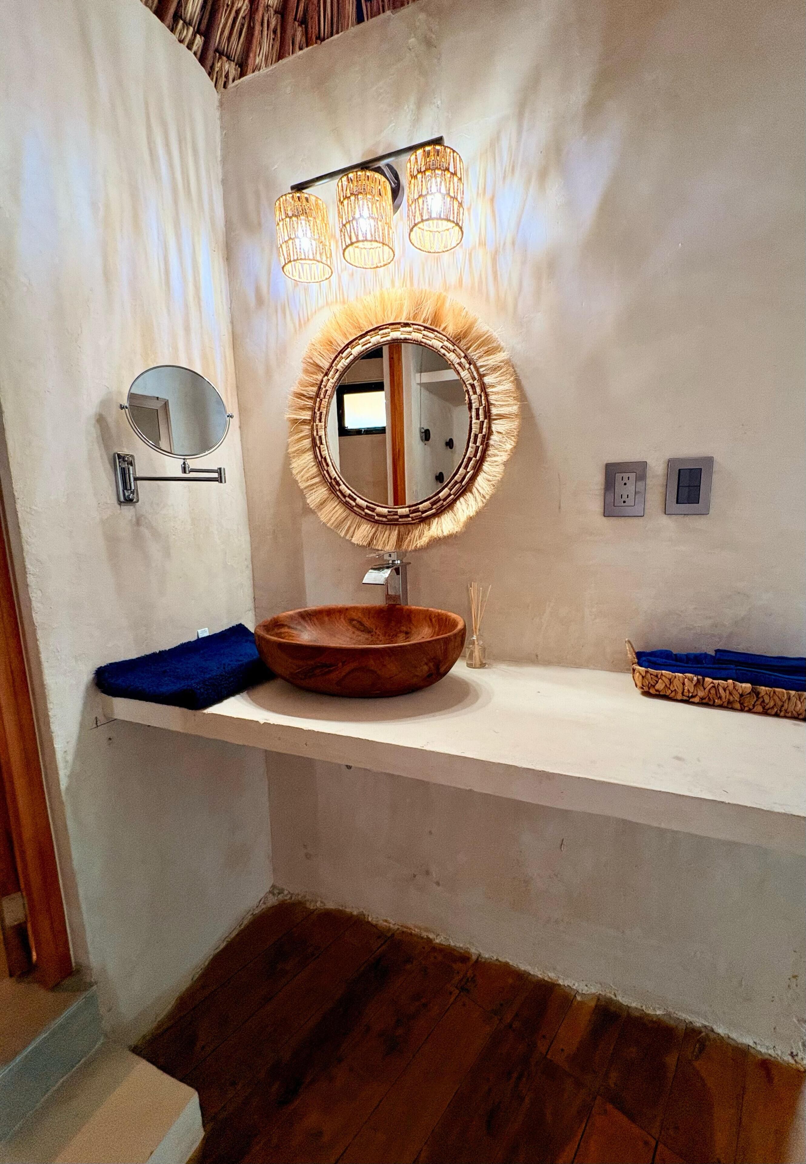 A bathroom with a wooden vanity, a round mirror with a woven frame, and a wall-mounted light fixture.