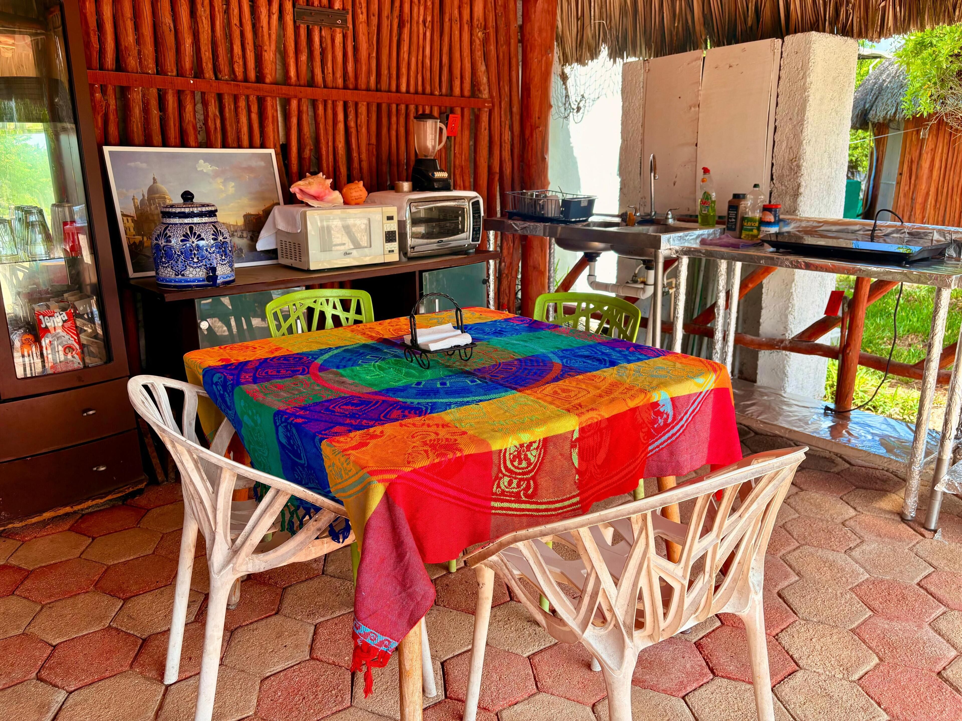 Een eethoek met een tafel, bedekt met een kleurrijke tafelkleed, witte stoelen, een magnetron, een broodrooster en een blender op het aanrecht.