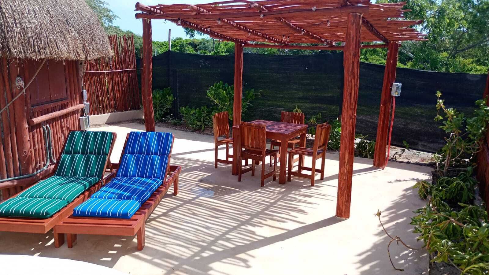 A wooden pergola with a table and chairs, a thatched-roof structure, and a fenced garden area.