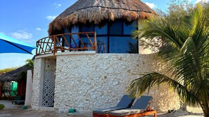Une villa située en bord de mer, dotée d’un toit de chaume, d’un mur en pierre et d’un petit balcon avec une balustrade en bois. On y voit deux chaises longues et un palmier au premier plan.