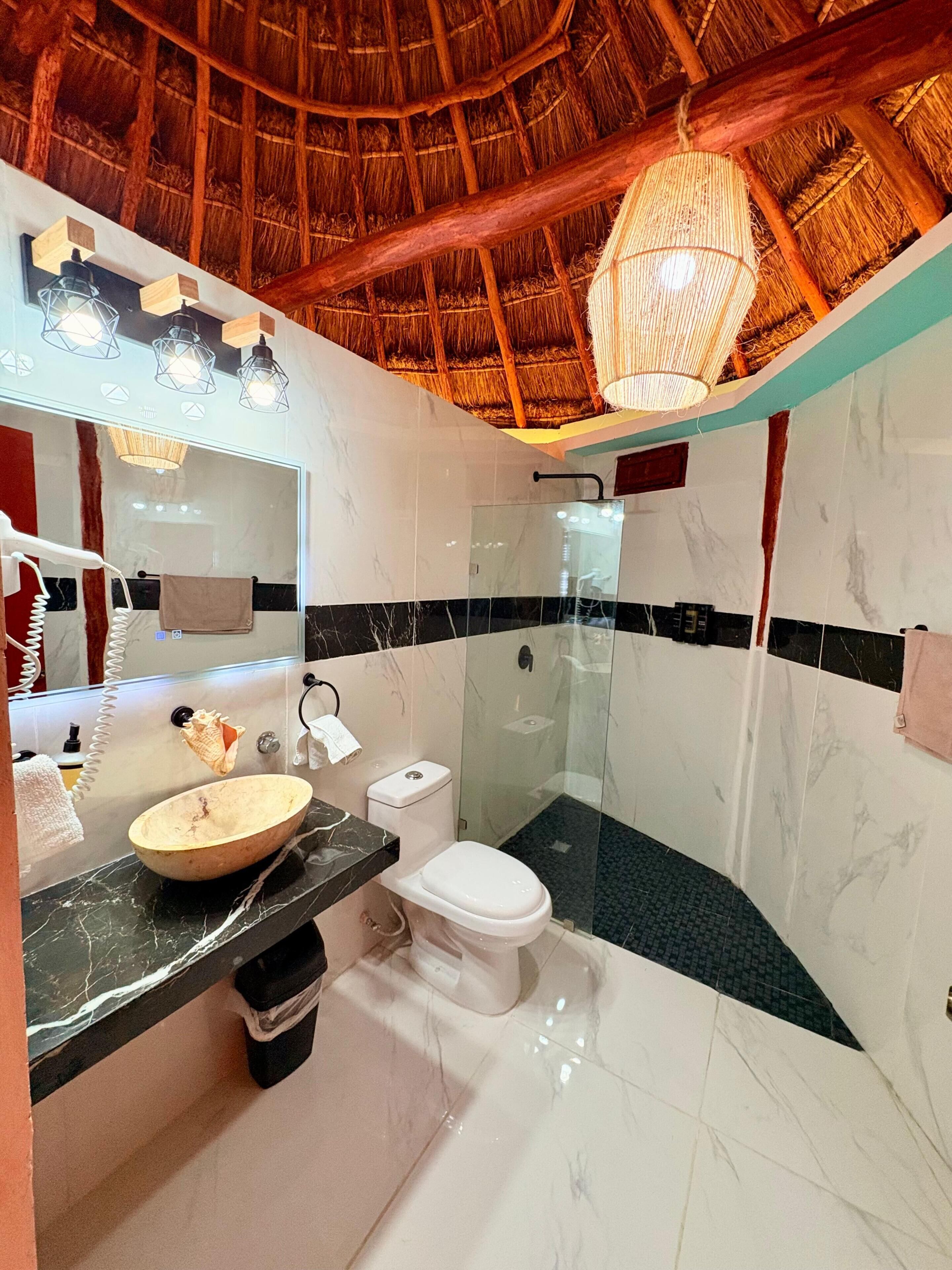 A bathroom with a glass-enclosed shower, a marble vanity, a white toilet, and a thatched ceiling.
