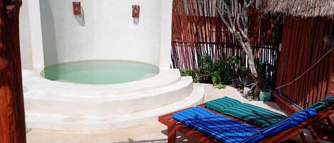 A hot tub with a wooden fence, a thatched roof structure, and a lounge chair with striped cushions.