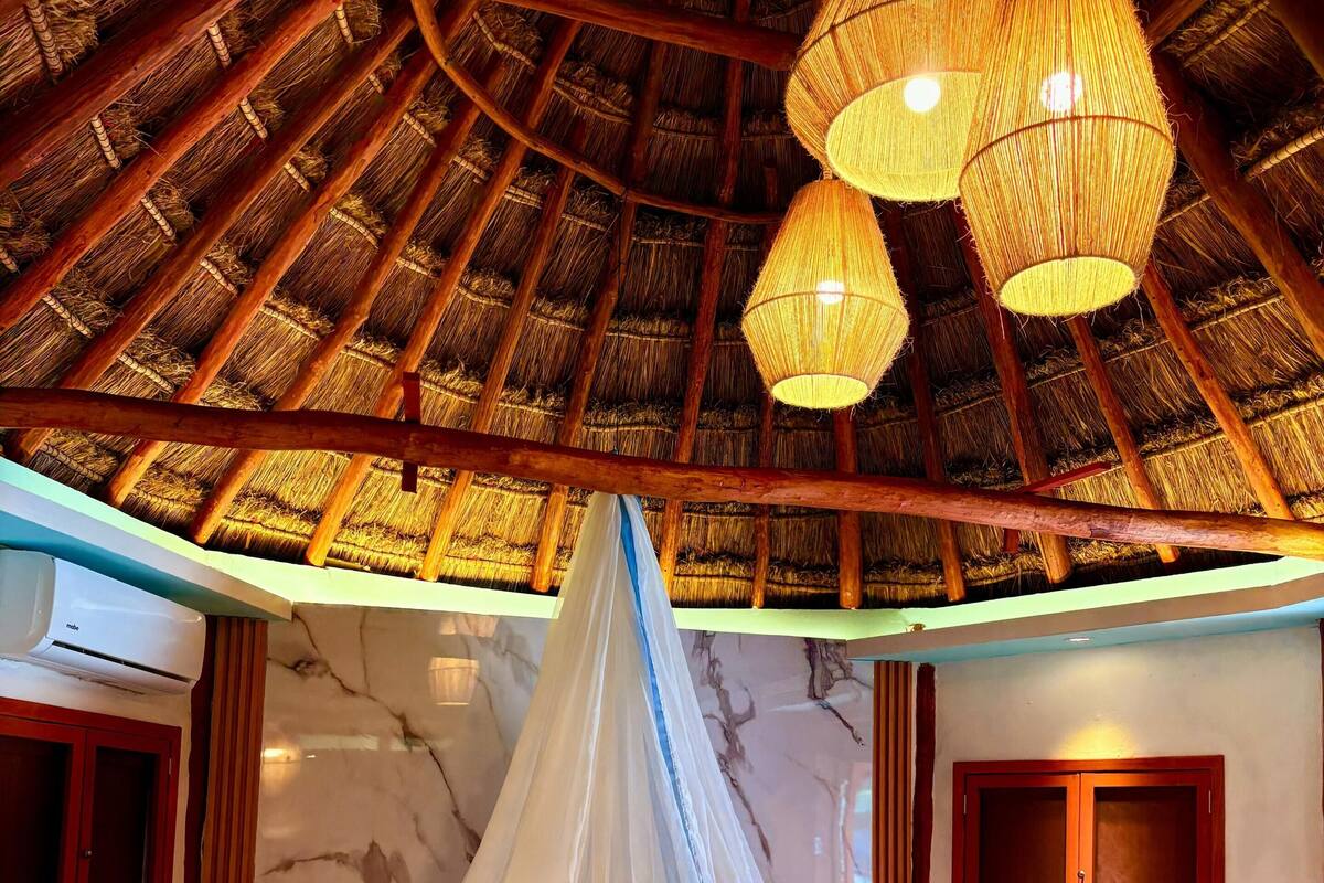 A room with a bed under a thatched roof, a hanging light fixture, and a wall-mounted air conditioning unit.