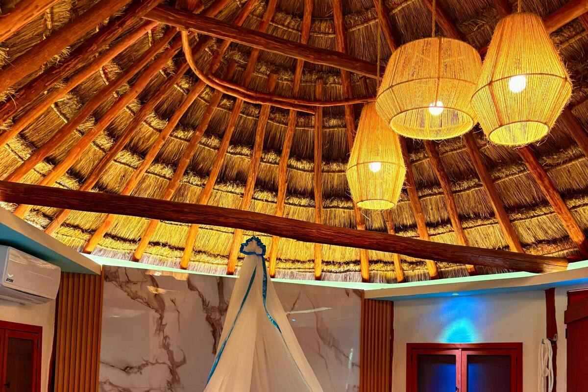 A room with a bed under a thatched roof, a hanging light fixture, and a marble wall.