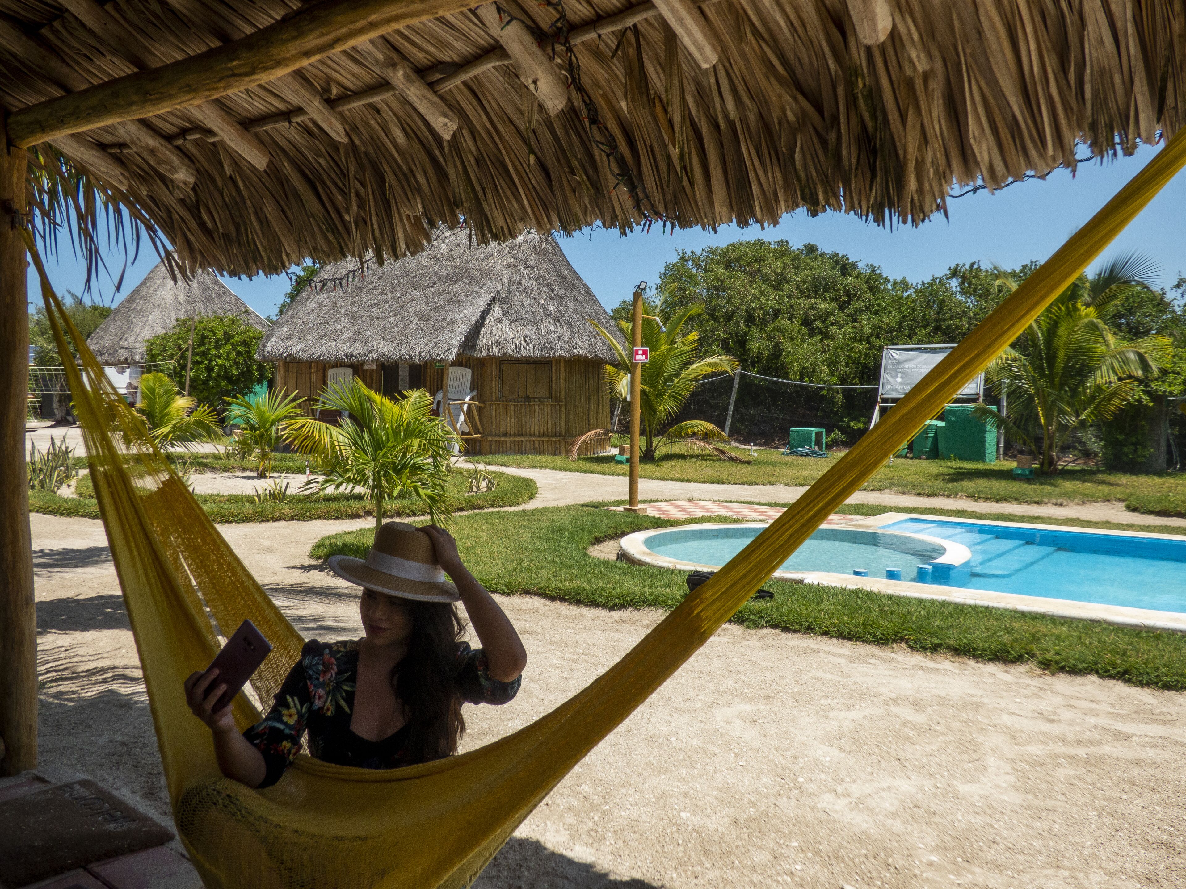 Una hamaca bajo techo de paja, una piscina y un edificio con techo de paja al fondo.