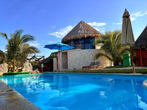A pool area with a thatched-roof building, lounge chairs, and palm trees.