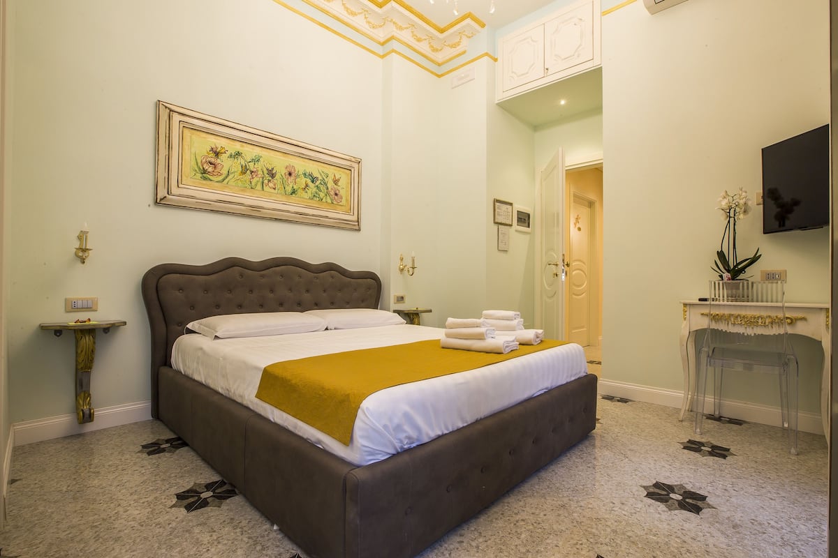 Una cama bien hecha con cabecera marrón, una obra de arte floral enmarcada encima y una mesita de noche pequeña con una lámpara.