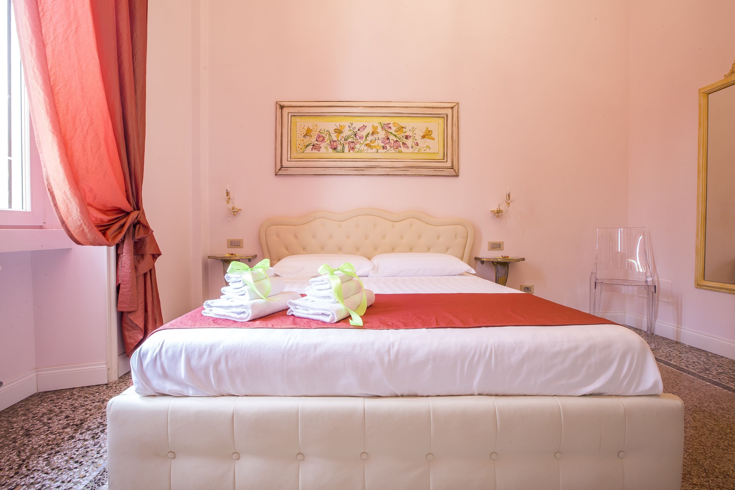 A neatly made bed with white and red linens, a framed floral artwork above it, and a clear glass chair to the side.