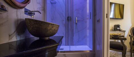 A bathroom with a marble sink, a large bowl, a shower with a glass door, and a television on the wall.