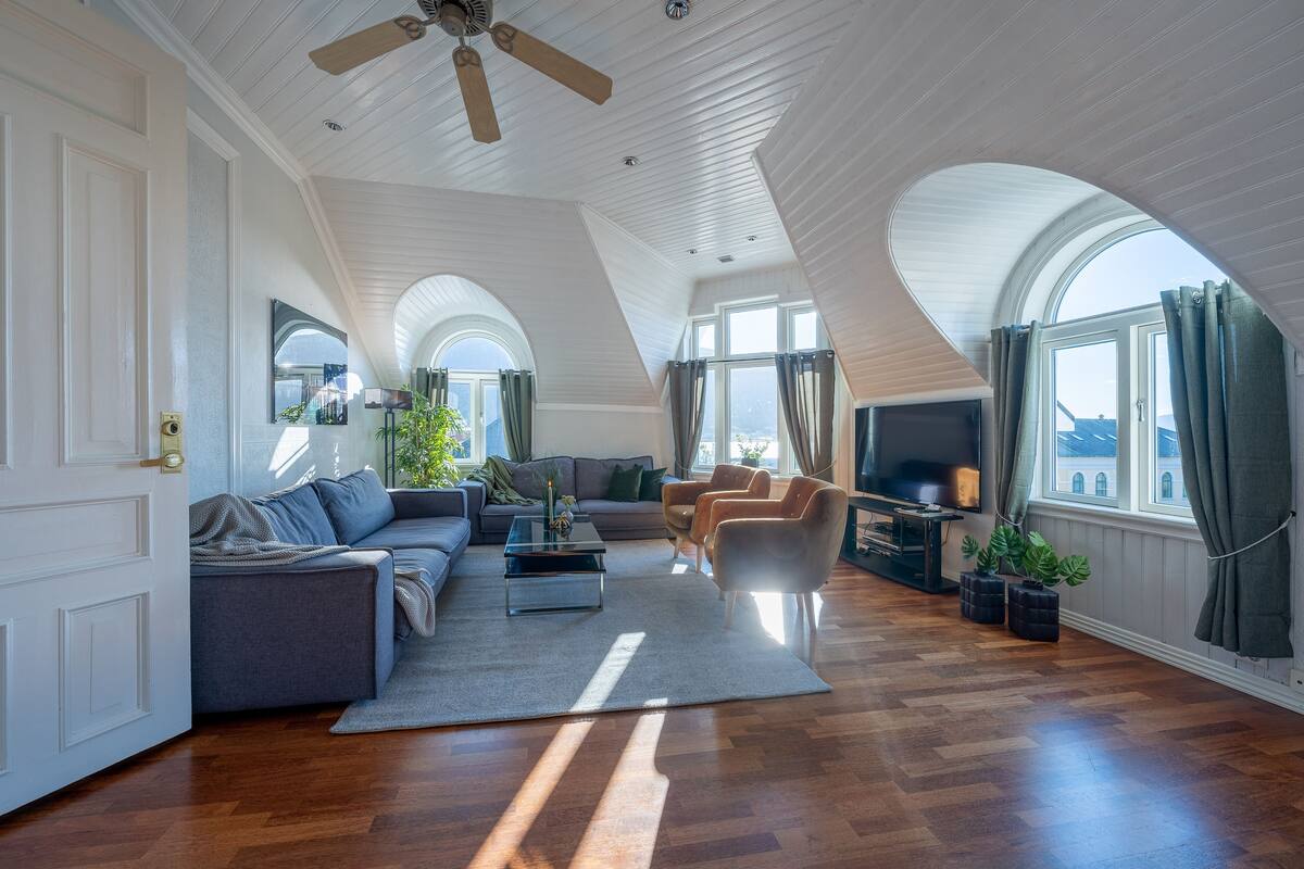 A spacious living room with a high ceiling, wooden flooring, a sofa set, a coffee table, and a television.