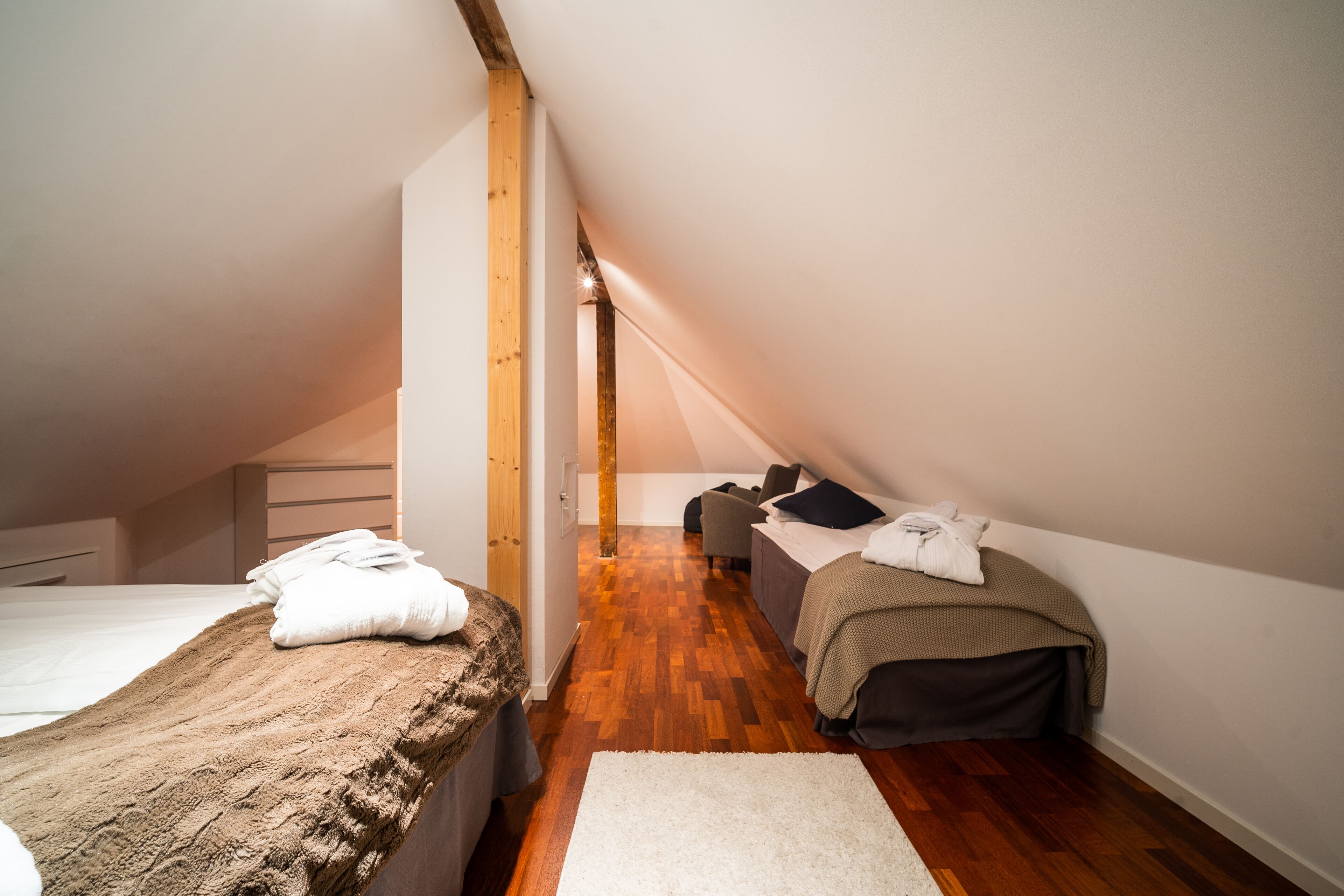 A bedroom with a sloped ceiling, wooden beams, a bed with a brown blanket, a bedside table, and a chair.