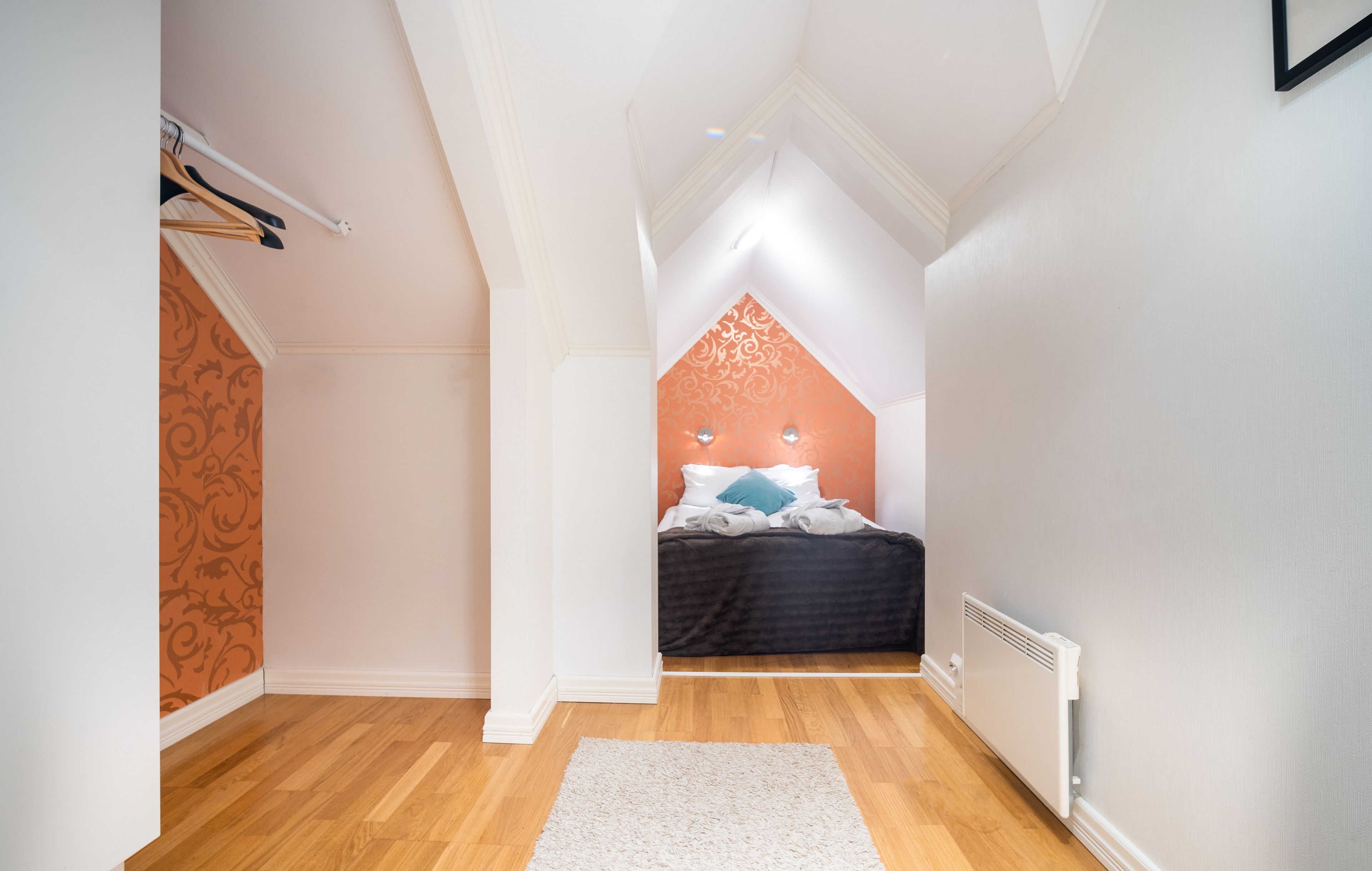 A bedroom with a sloped ceiling, a bed with white and blue bedding, a radiator, and a patterned wall.