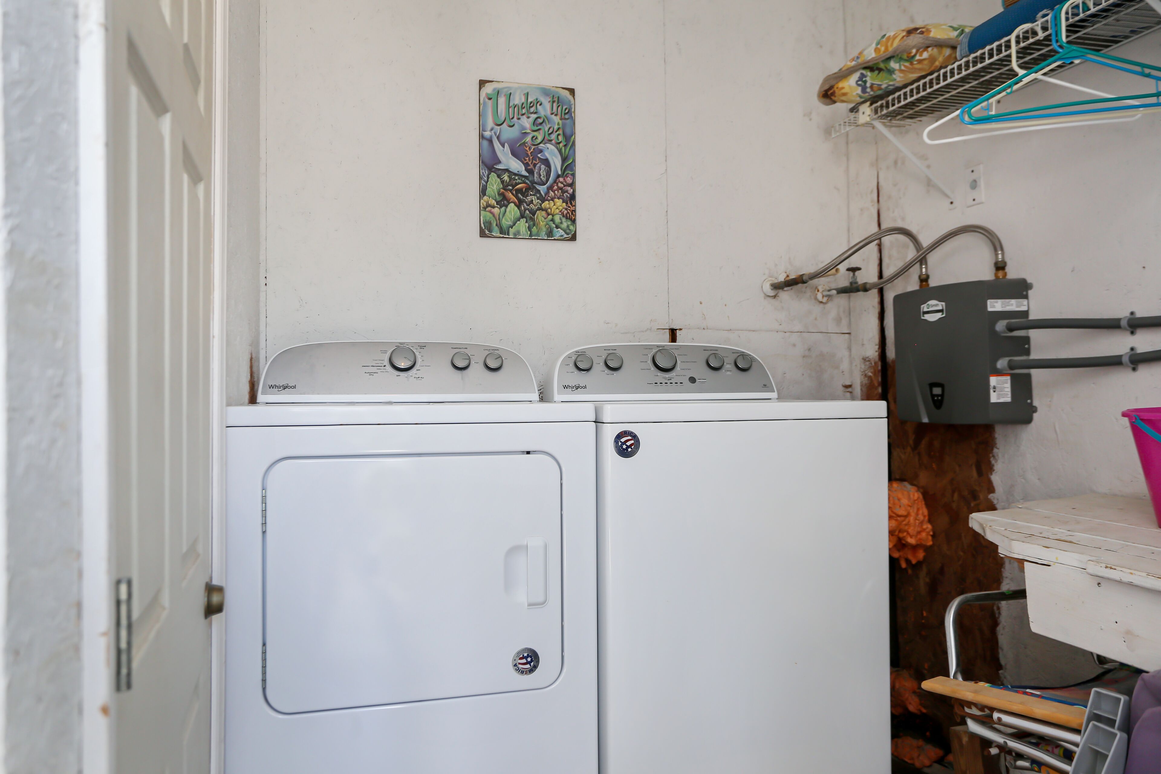 Un espace buanderie compact comprenant un lave-linge et un sèche-linge blancs, un étendoir métallique avec des vêtements et un chauffe-eau fixé au mur.