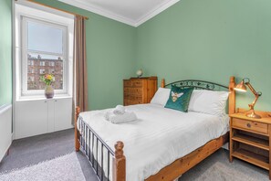 A bedroom with a bed, a nightstand, a dresser, a window with curtains, and a view of buildings outside.