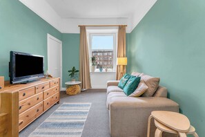 A room with a television, a sofa, a small table, a window with curtains, and a view of buildings outside. - Bright Quiet Edinburgh Home Free Parking (Newhaven)