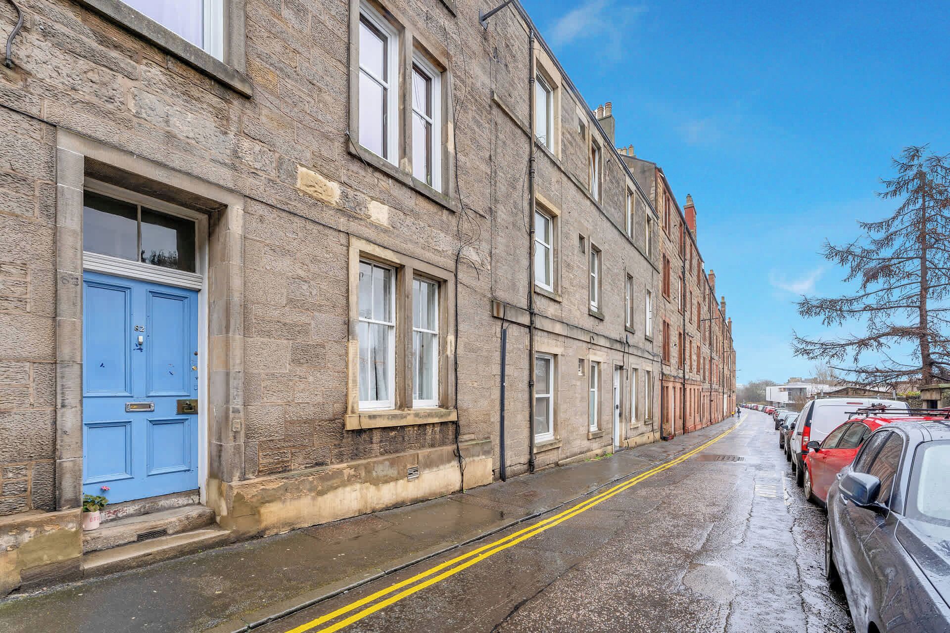 A row of traditional stone buildings with a blue door on the left, parked cars on the right, and a clear sky.
