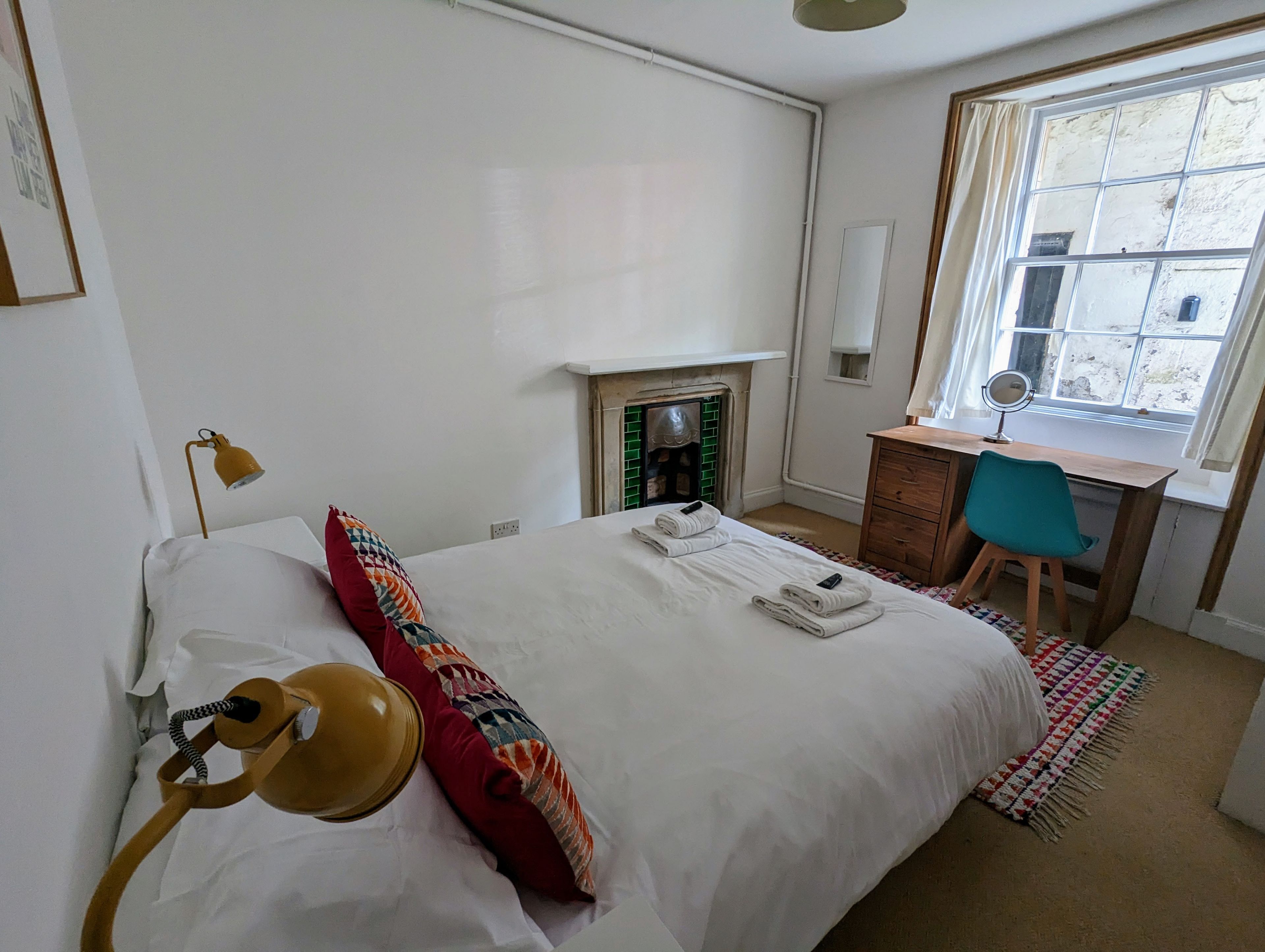 A bedroom with a bed, a desk, a chair, a lamp, a fireplace, and a window.