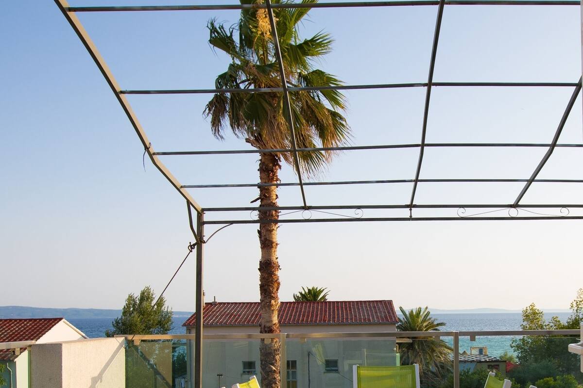 A terrace with a white table and green chairs, overlooking a coastal area with a palm tree and buildings in the background.