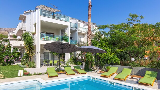 Un hotel moderno con piscina, sillones reclinables verdes y un cielo azul despejado.