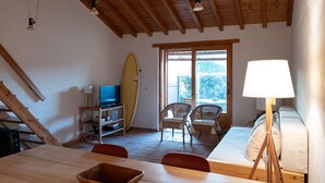 Ein gemütliches Zimmer mit Holzbalkendecke, einem an die Wand gelehnten Surfbrett, einem Fernseher, einem Esstisch mit Stühlen und einem Bett mit Lampe.