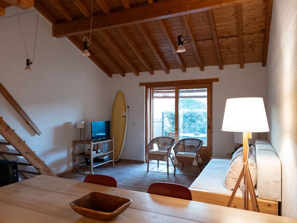 Une pièce chaleureuse avec un plafond en bois, une planche de surf appuyée contre le mur, une télévision, une table à manger avec des chaises et un lit avec une lampe.