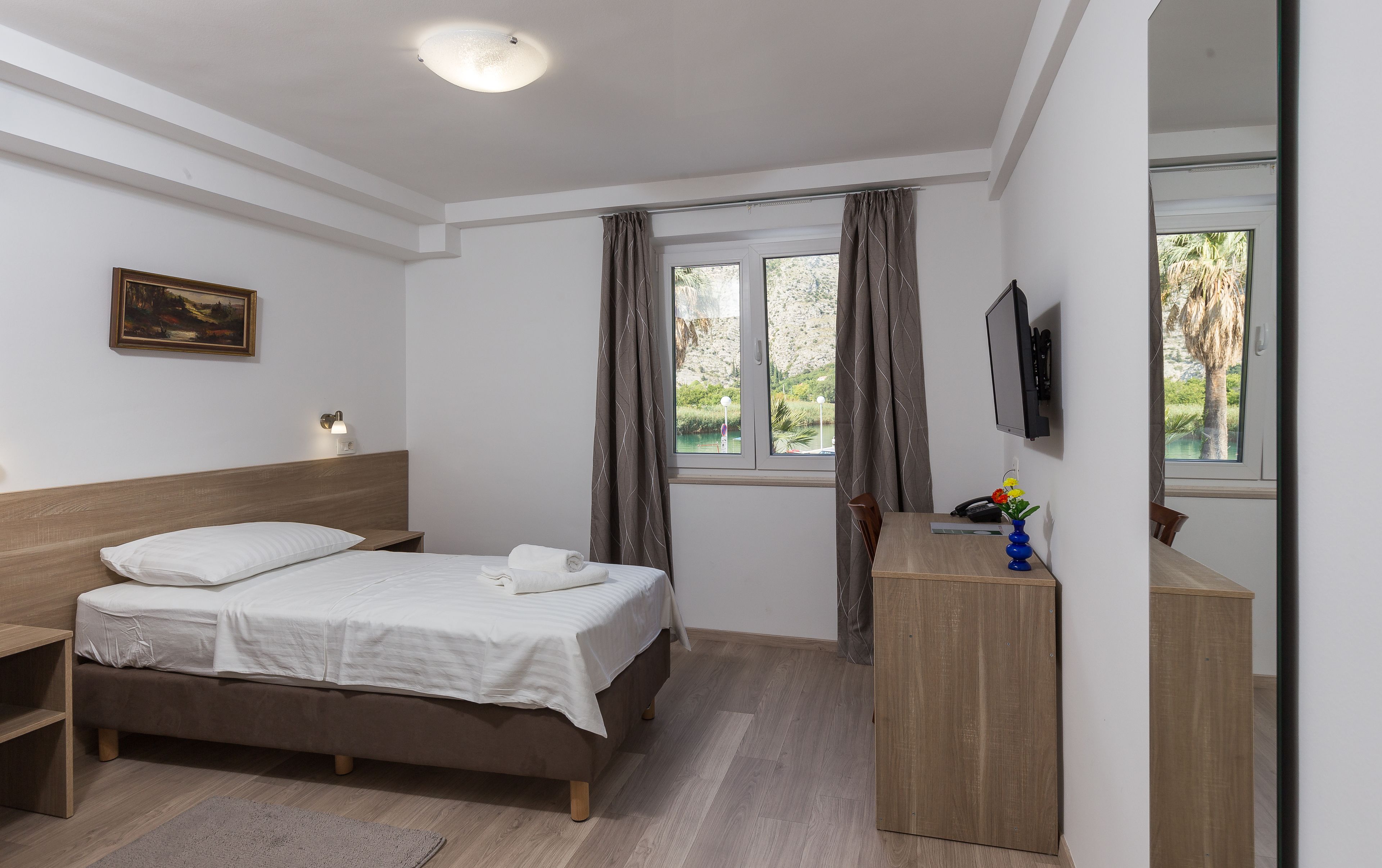 A bedroom with a bed, bedside table, a desk, a chair, a TV mounted on the wall, and a window with curtains.
