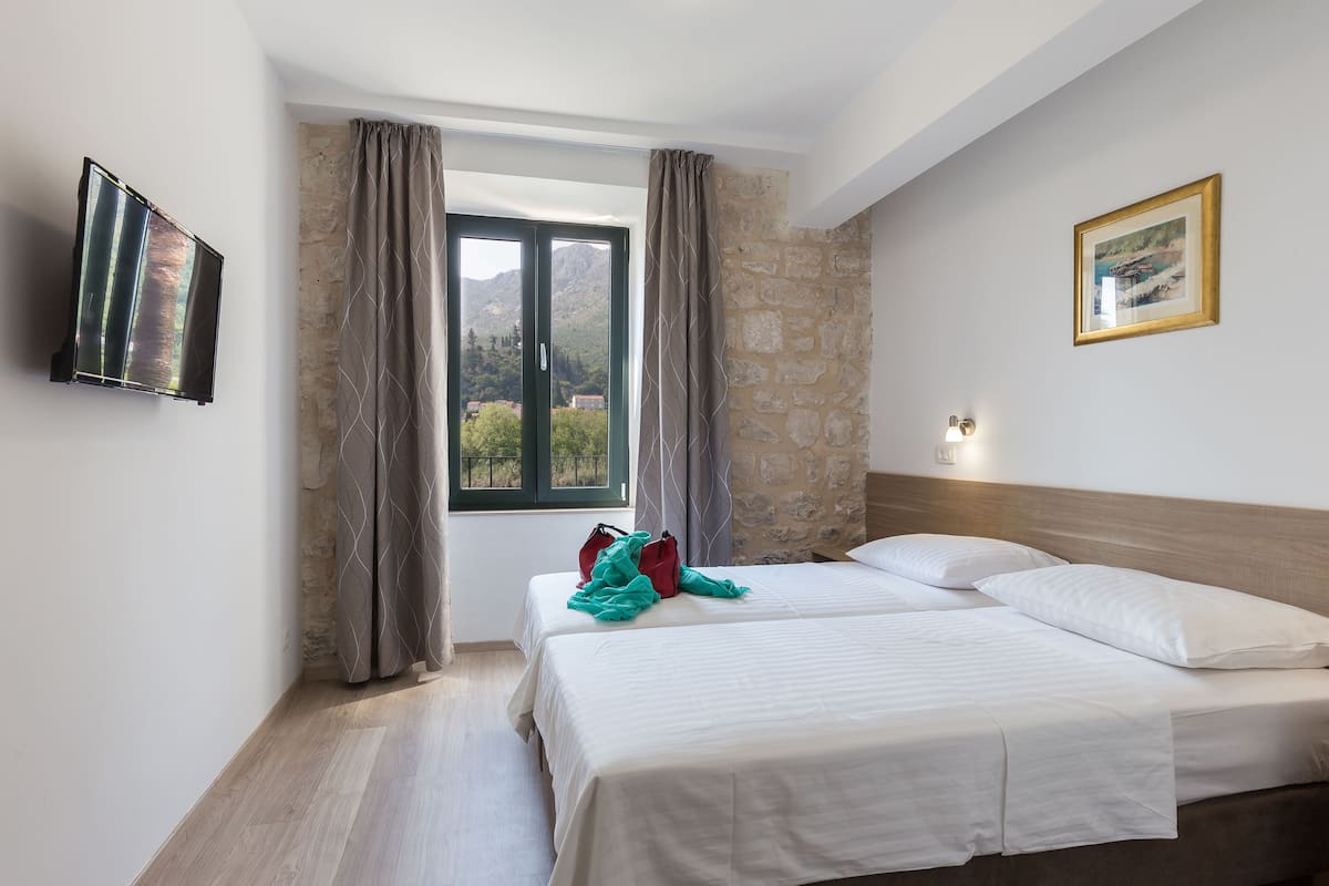 A hotel room with two beds, a TV, a window with curtains, a stone wall, and a framed picture on the wall.