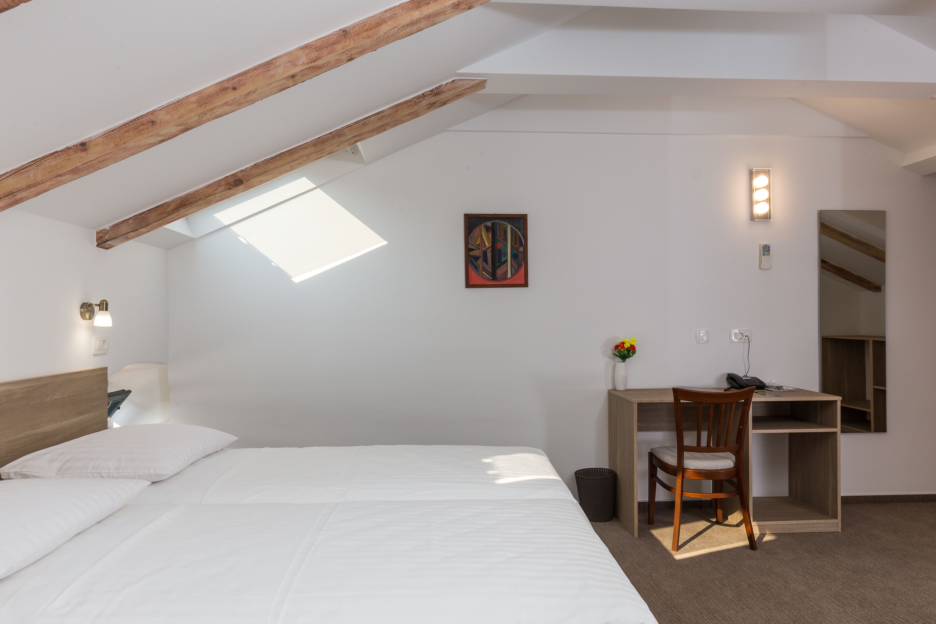 A bedroom with a bed, a desk, a chair, a small trash bin, a wall-mounted light, and a skylight.
