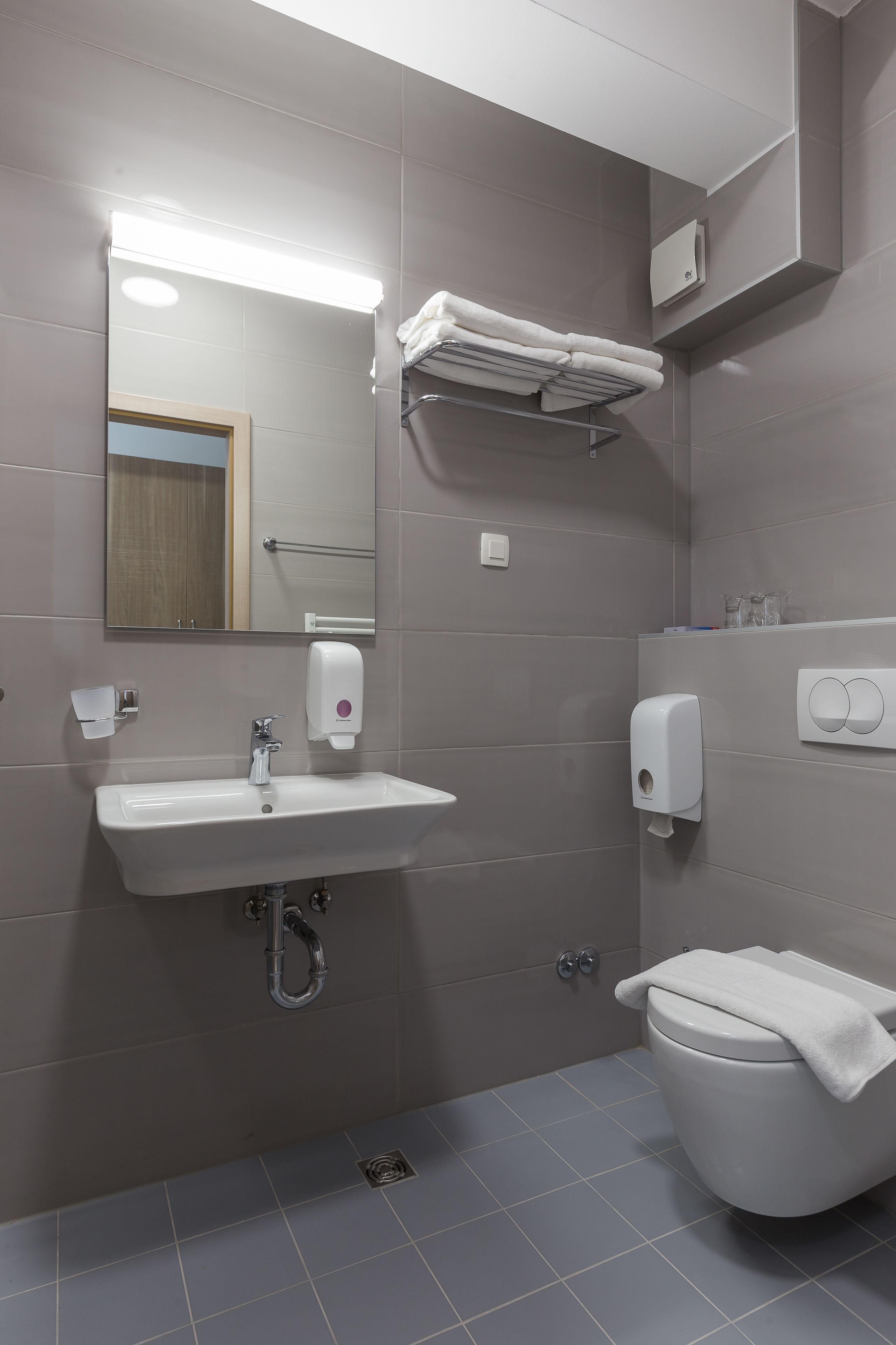 A modern bathroom with a white sink, mirror, and toilet. There are towel racks and a soap dispenser on the wall.