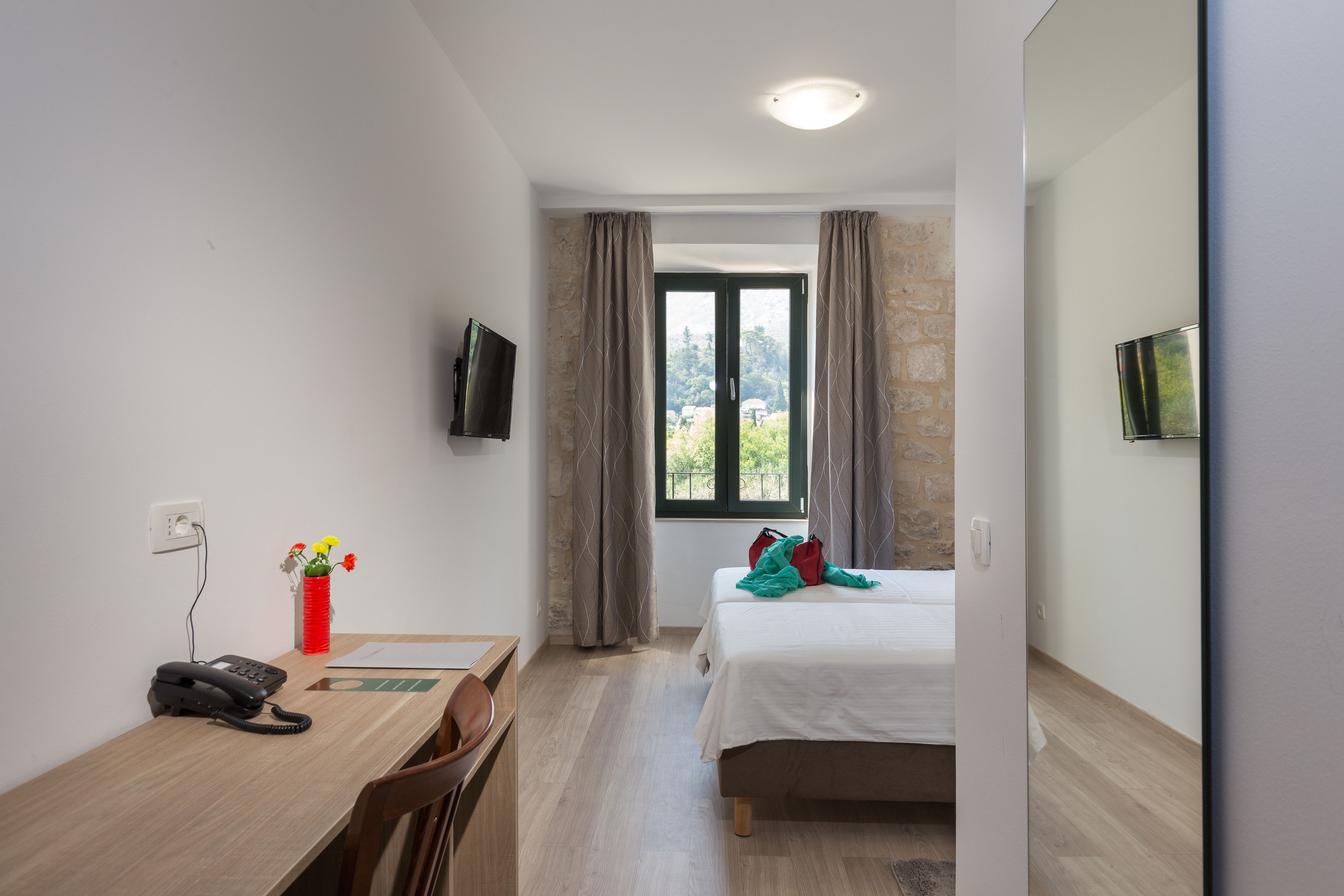 A hotel room with a bed, a desk with a telephone, a chair, a TV mounted on the wall, and a window with curtains.
