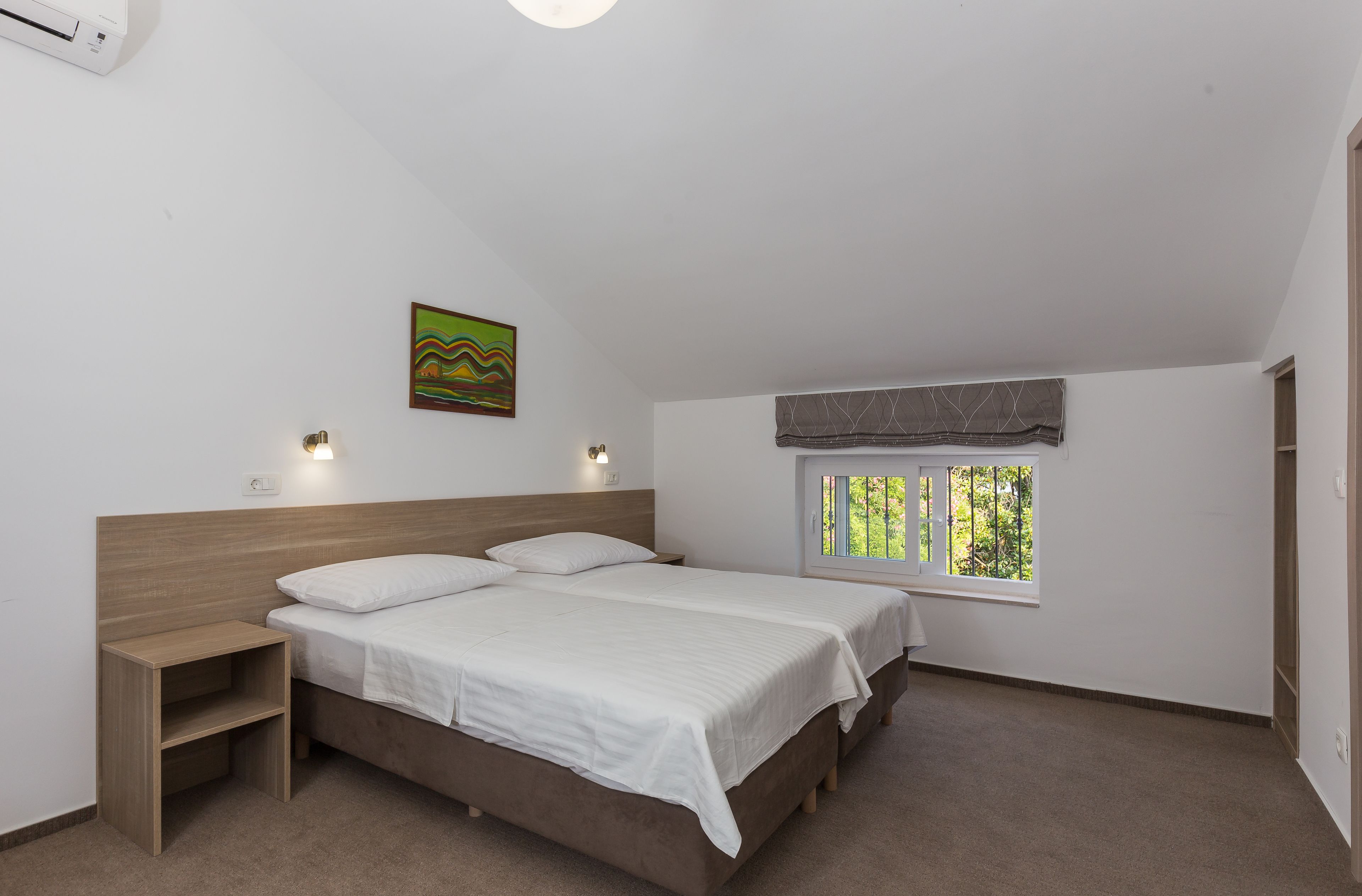 Un dormitorio con cama, mesitas de noche, ventana con vistas y aire acondicionado instalado en la pared.