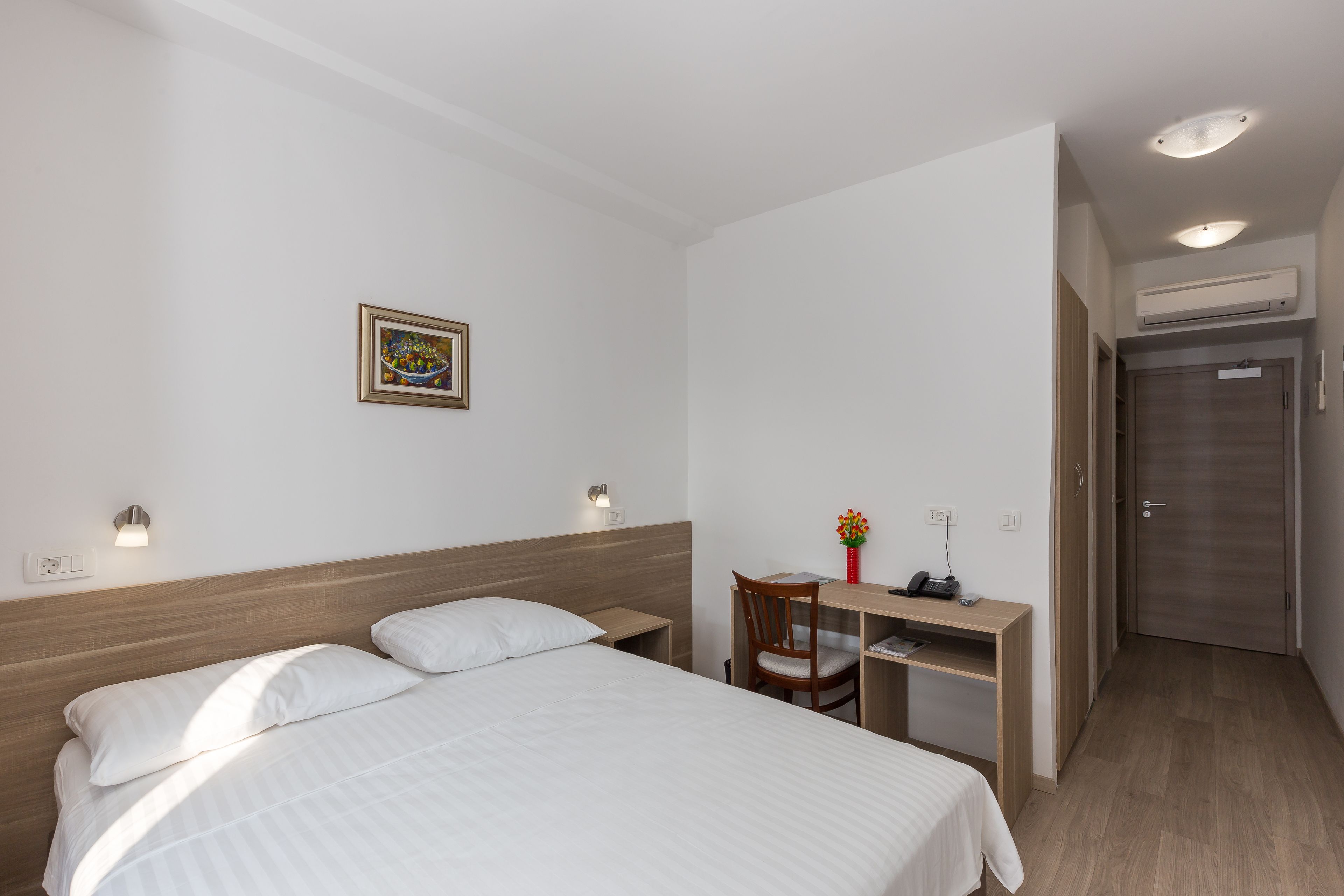 A hotel room with a bed, a desk, a chair, a small table, a wall-mounted shelf, a framed picture, and an air conditioning unit.