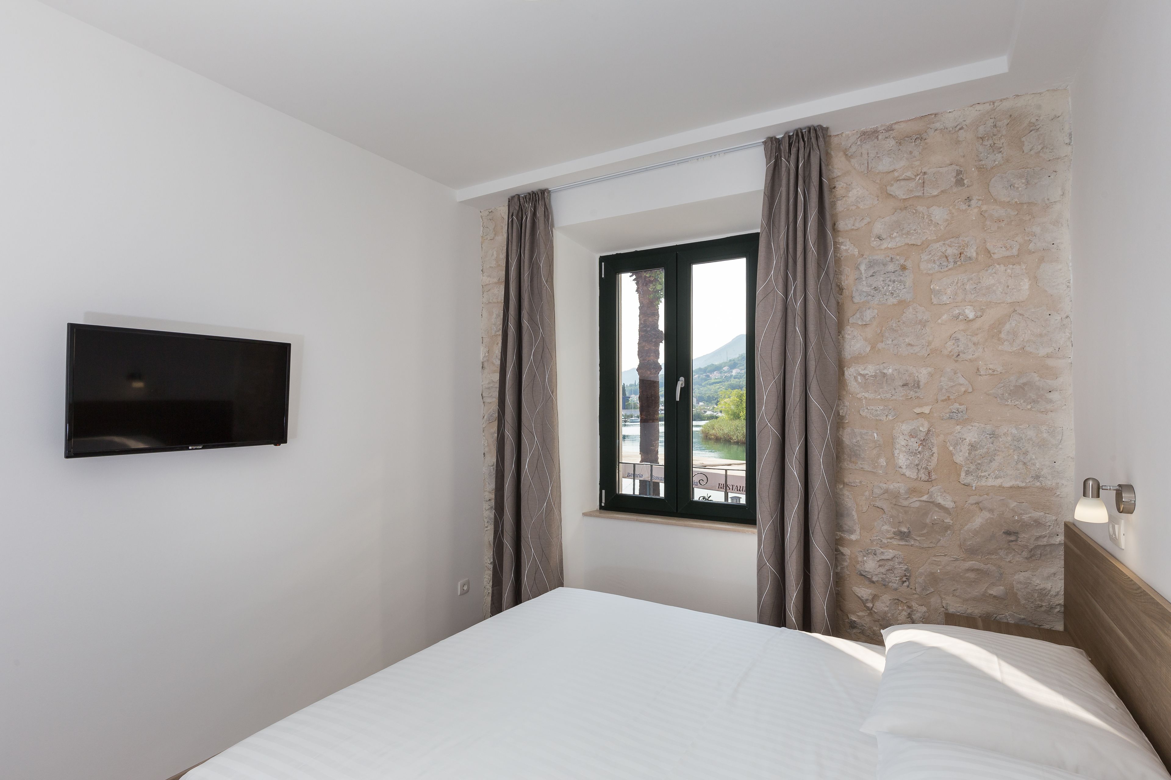 A hotel room with a bed, a TV mounted on the wall, a window with curtains, and a stone wall.