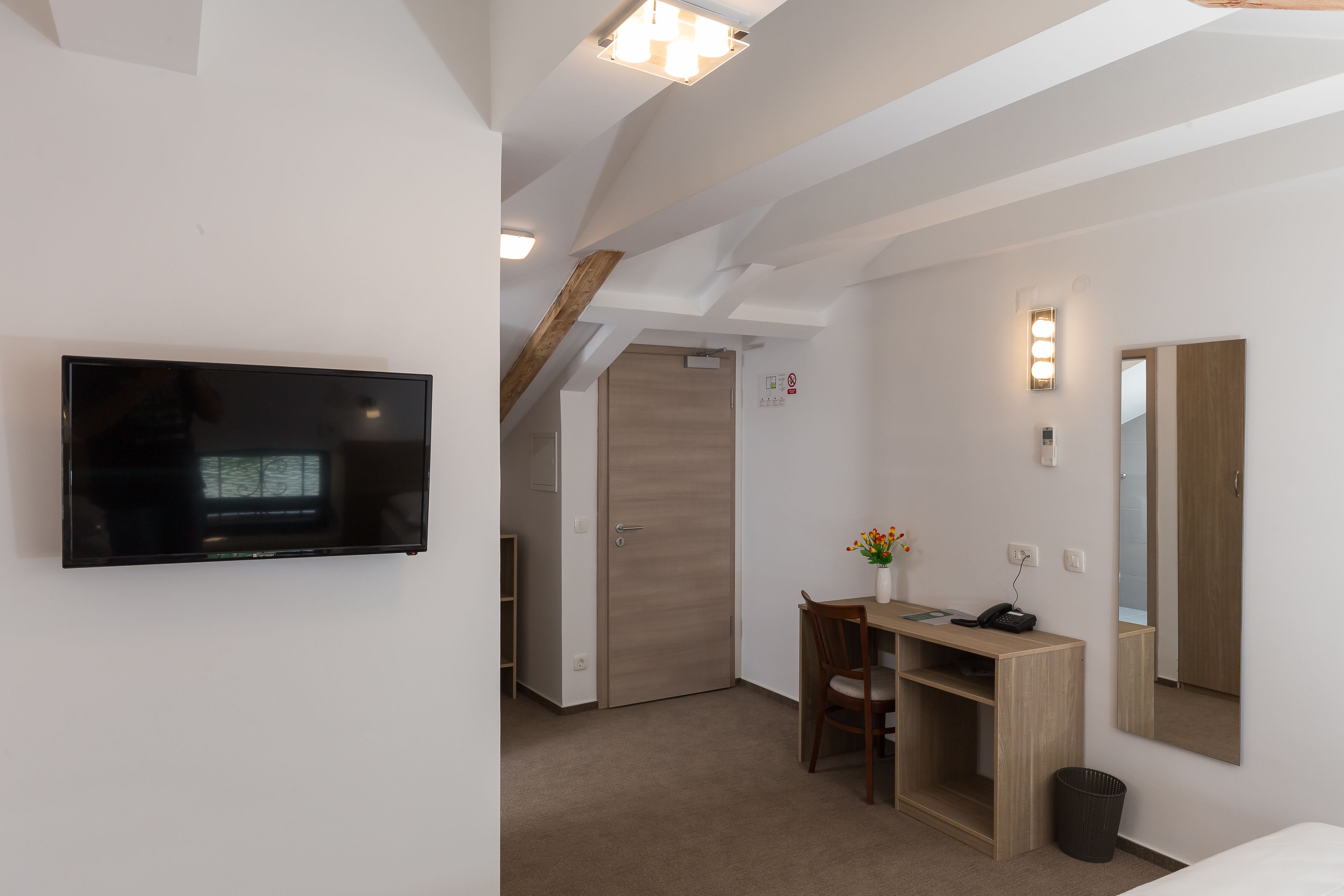 Ein Hotelzimmer mit einem an der Wand befestigten Flachbildfernseher, einem Schreibtisch mit Stuhl, einer kleinen Mülleimer und einem Wandspiegel.